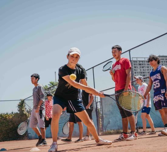 Plano general de mujer sosteniendo una raqueta de tenis a punto de pegarle a una pelota en el aire en una cancha. Detrás de ella hay siete personas más con raquetas de tenis en las manos.