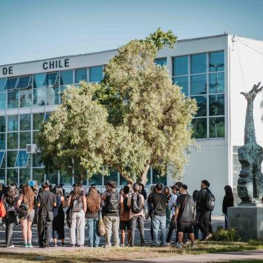 Grupo de estudiantes frente a la Casa Central de la Universidad. Al costado del estudiantado se encuentra la escultura en memoria de Victor Jara, una guitarra que se fusiona y finaliza con una mano a lo largo de su estructura.