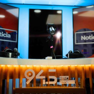 Radio Usach de la Universidad. En la imagen aparecen tres pantallas verticales con el texto "Noticias" en las pantallas externas. Frente a ellas se ven tres micrófonos de estudio sobre un mesón y debajo de este se encuentra el logo impreso "94.5 Radio Usach" en color blanco.