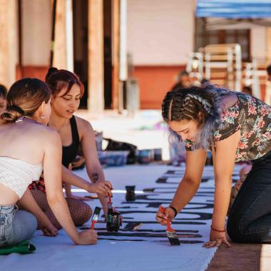 Tres mujeres pintando una consigna en un lienzo blanco con brochas y pintura negra en el piso de la Escuela de Artes y Oficios.