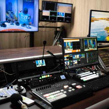 Sala de control de Stgo.TV, canal de televisión de la Usach. En la foto aparecen dos pantallas de computador junto a una consola, las cuales están sobre un mesón. Frente a ellas se ubican dos pantallas de televisión colgadas.