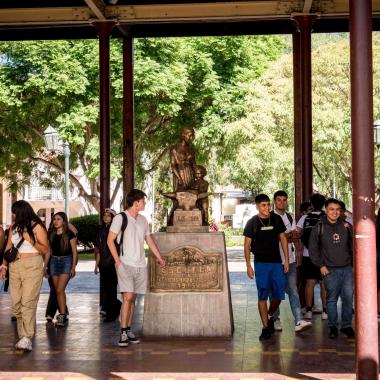 Estudiantes caminando por el pasillo central de la Escuela de Artes y Oficios. La escultura "Alma Mater" se encuentra en medio de este pasillo y de la gente. 