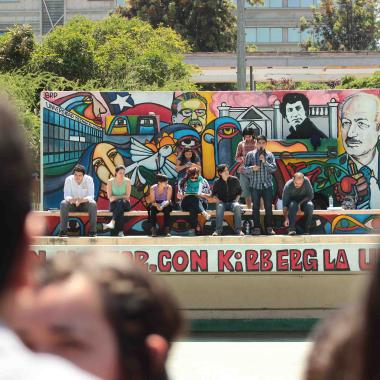 Foro griego de la Universidad. Se enfoca el escenario pequeño que contiene un mural con personajes y espacios icónicos de la comunidad usachina: Salvador Allende, Victor Jara y Enrique Kirberg. También está ilustrada la Universidad Técnica del Estado, nombre que llevaba la Universidad de Santiago de Chile antes del 1981, plena dictadura en Chile. Frente a este mural sobre el escenario se encuentran seis personas sentadas y dos de pie. Una de las personas paradas se encuentra con un micrófono hablando. 