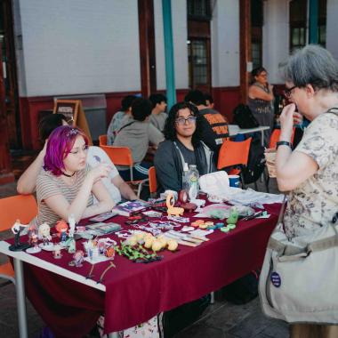 Dos estudiantes sentadas en una mesa en uno de los patios de la Escuela de Artes y Oficios vendiendo collares, llaveros, espejos, etc 