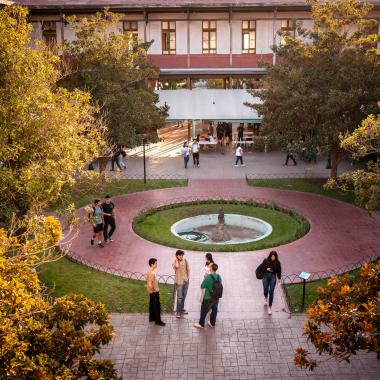 Patio de la EAO enfocado desde el segundo piso. La vista apunta hacia la pileta central que posee una escultura dentro. Alrededor de la pileta se encuentran estudiantes conversando.