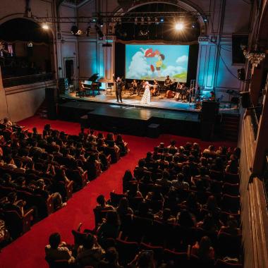 Interior del Aula Magna visto desde el segundo piso. En el escenario aparece una mujer y un hombre cantando junto a la agrupación musical ubicada detrás. Se encuentran interpretando un concierto sobre la música popular del Studio Ghibli y al fondo se puede apreciar un fotograma de una de las películas producidas por ese estudio. Al frente del escenario están todos los asientos ocupados por la audiencia.