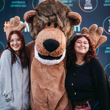 Dos chicas posando felices con la mascota de la Universidad en medio de ellas. La mascota, conocido como Usachín, es una persona disfrazada de león, personaje que representa a las y los estudiantes de la universidad.