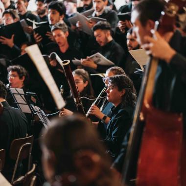 La cámara enfoca a una joven que está de perfil tocando un instrumento de viento. A sus costados le acompañan personas cantando y otras tocando instrumentos de cuerda y viento.