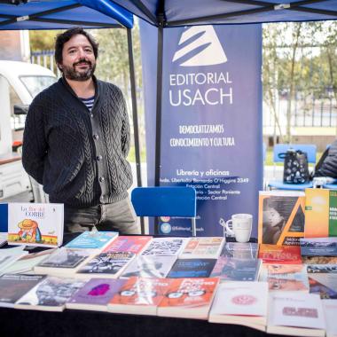 Persona sonriente atiende el puesto de libros de la Editorial Usach en el contexto de una feria de lectura en la universidad. Detrás de la persona se ubica un pendón azul indicando el nombre de la editorial. 