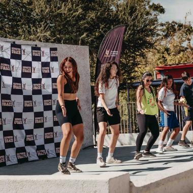 Plano general enfocado desde un costado de un escenario. Aparecen cinco guías practicando zumba sobre el escenario del foro griego del campus universitario.