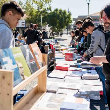 Estudiantes y funcionariado revisan libros que están instalados en mesones a lo largo de la entrada de la universidad.