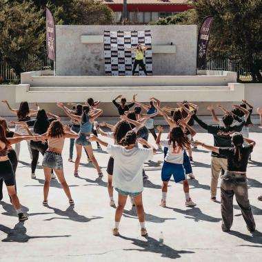 Plano general del foro griego de la universidad. Sobre el escenario que está al fondo hay una profesora guía de zumba. Al frente de ella hay veinticuatro jóvenes siguiendo sus pasos de espaldas a la cámara.