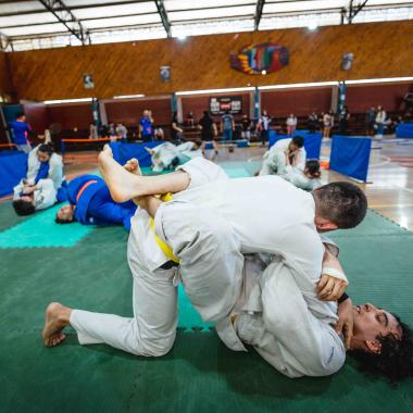 Plano entero de dos chicos practicando judo. Realizan una posición de llave en el suelo, mientras que detrás hay cuatro duplas más practicando el deporte sobre unas colchonetas. Más atrás se ven otras personas practicando otros deportes. 