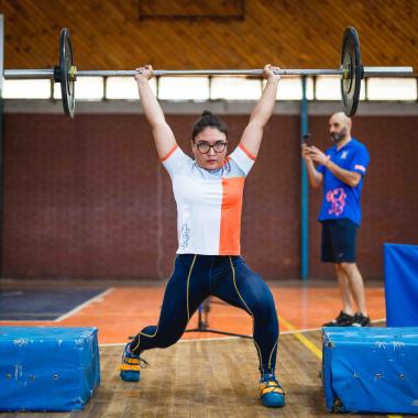 Plano entero en un gimnasio de una chica levantando una pesa con una rodilla más flectada que la otra.