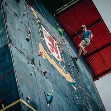 Plano general de un joven sostenido por una cuerda. Está mirando hacia abajo mientras desciende de escalada. En el muro que está descendiendo se encuentra pintado el escudo de la universidad.