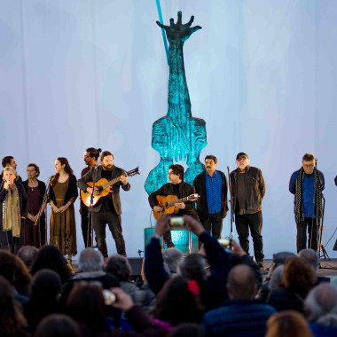 Grupo de artistas chilenos y chilenas sobre un escenario en la universidad: Isabel Parra, Manuel Meriño, Horacio Salinas, Cristián Mancilla, José Seves (Inti-Illimani Histórico), Cuncumén, Ismael Oddó y Nano Stern. Detrás aparece la escultura en memoria de Victor Jara iluminada. 