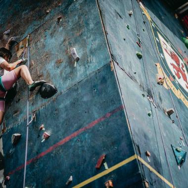 La imagen enfoca un muro de escalada que tiene la forma de un prisma. En su costado izquierdo una chica está escalando hacia arriba, mientras que en la otra cara del muro un joven lo está descendiendo. A ambos les sostiene una cuerda.