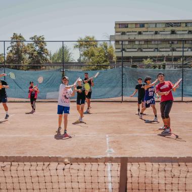 Plano general de una cancha de tenis. Al medio de la cancha hay cuatro columnas conformadas de tres jóvenes cada una. Quienes están ahí sostienen una raqueta de tenis a modo de práctica.