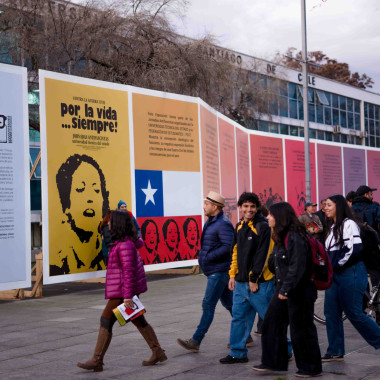 Exposición "Por la vida... ¡siempre! (1973-2023)" en el frontis de la Casa Central de la Universidad. Son paneles de gigantografías coloridas de afiches creados el año 1973 con llamados para evitar la guerra civil de la época. Delante de esta exposición se encuentran personas caminado y observando. 