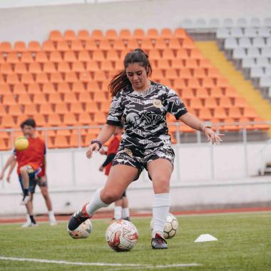 Plano entero de una joven jugando futbol en una cancha con pasto. Detrás de ella se ven dos muchachos y los asientos de las galerías.