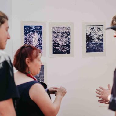 Aparecen dos jóvenes viendo hacia el frente cómo una señora está pegando cuatro impresiones de xilografía en una pared para montar una exposición.