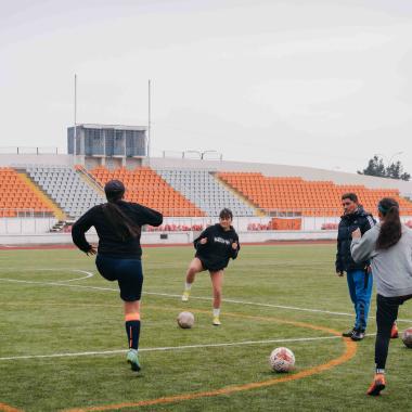 Plano general de cinco jóvenes en círculo y un entrenador realizando precalentamiento para practicar fútbol. Detrás se puede ver los asientos de la galería de la cancha.