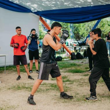 Plano entero de dos jóvenes practicando boxeo en el pasto al interior del campus universitario. Detrás aparecen cinco jóvenes con guantes de boxeo en las manos viendo a la dupla. 