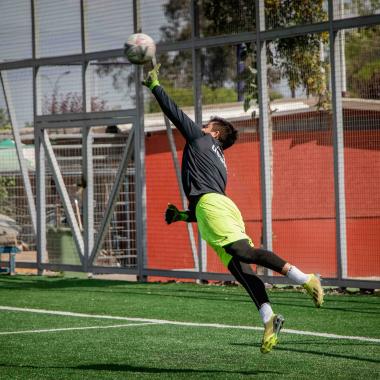 Plano entero de arquero atrapando una pelota de fútbol mientras salta a su costado derecho. 