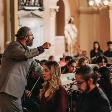 Un director de orquesta se encuentra de perfil en el lado izquierdo de la foto dirigiendo a las y los músicas(os), quienes se encuentran sentadas y sentados tocando sus respectivos instrumentos.