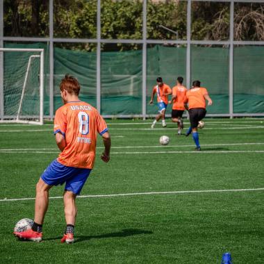 Plano general de un joven de espalda jugando fútbol y al frente de él hay cuatro jóvenes más jugando cerca del arco de la cancha.