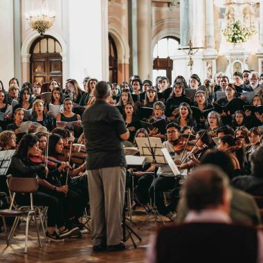 Persona dirige orquesta musical. En escena aparecen violinistas sentadas y sentados frente al director. Detrás de ellas y ellos les acompaña un coro.