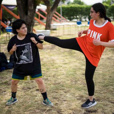 Plano entero de dos muchachas practicando defensa personal. Una de las chicas mira a la otra esperando un ataque con los brazos apegados a su pecho y la otra chica está de perfil levantando su pie derecho hasta el hombro de la otra muchacha.