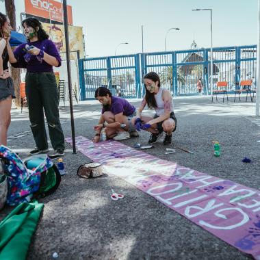 se muestran cuatro personas, donde 2 estan conversando y las otras pintan un lienzo en conmemoracion al dia de la mujer