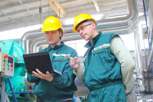 Dos profesionales se desenvuelven en el laboratorio. Trajes verde petróleo, cascos amarillos.