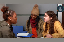 Foto de personas migrantes compartiendo en clase. Mujer con gorrito. Joven con chaleco café claro y otra con chaqueta.