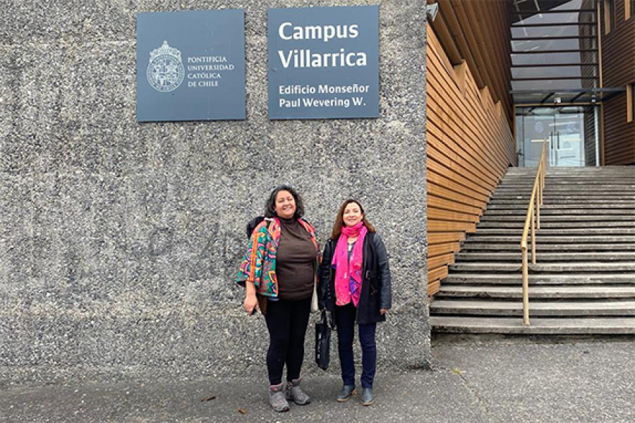 Dos mujeres posan en muralla de una universidad. Al costado aparece una escalera