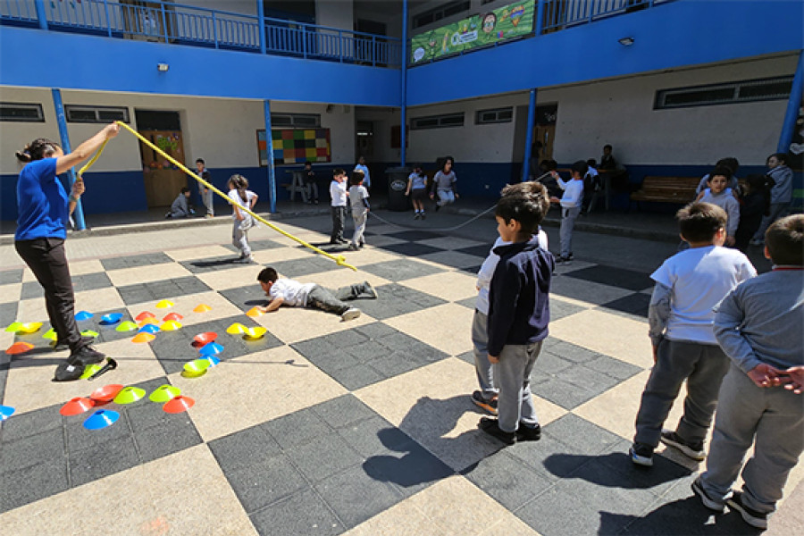 Niños juegan con su profesor en e patio de la escuela. Suelo como tablero de ajedrez y fondo en azul y blanco.