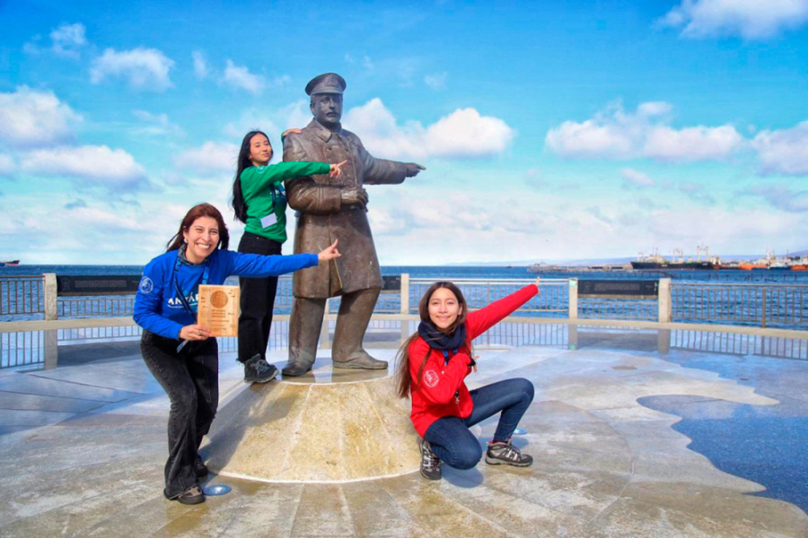 Claudia Mariela Pizarro Ojeda, profesora de Química y Ciencias Naturales,  junto a las estudiantes Antonia Acuña y Antonia Guerrero en Punta Arena. En la fotografía se logra ver a la profesora, las dos estudiantes, el estrecho de Magallanes y una estatua que apunta hacia la Antártica