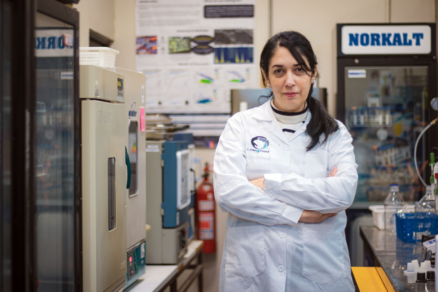 Dra. Jhosané Pagés, investigadora del Departamento de Ingeniería Química y Bioprocesos., posa a la cámara en su laboratorio de investigación. Lleva puesta un delantal blanco y los brazos cruzados.