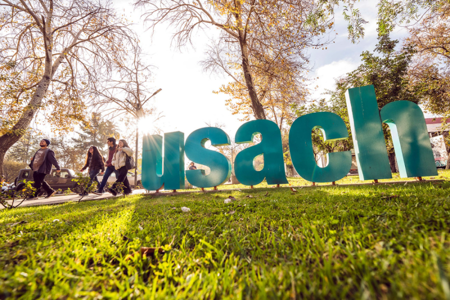 Letras institucionales al ingreso de la Universidad por metro Usach. Son de tamaño gigante en color verde y las letras están sobre el pasto del Campus. Por su lado izquierdo caminan 4 estudiantes.