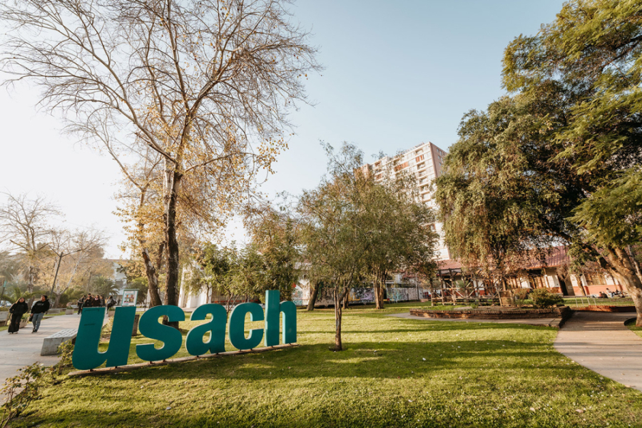 En la imagen aparecen las letras gigantes que indican USACH. Éstas, están instaladas en la entrada por el Departamento de Educación y el Departamento de Filosofía, cercano al metro Usach. En la imagen se ven los pastos y las instalaciones correspondiendo a corredores coloniales de la Universidad y a tres estudiantes que caminan hacia la reja principal.