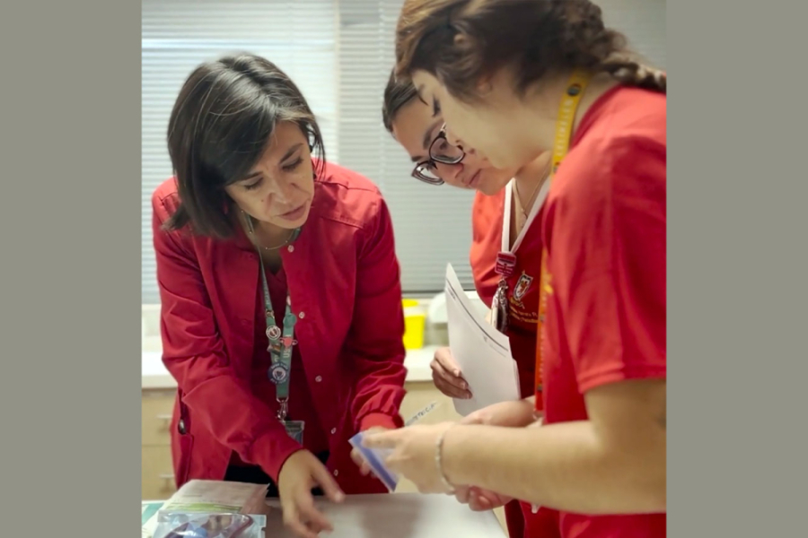Académica enseña a dos estudiantes de Ciencias Médicas. Todas vistes ropajes en color rojo.