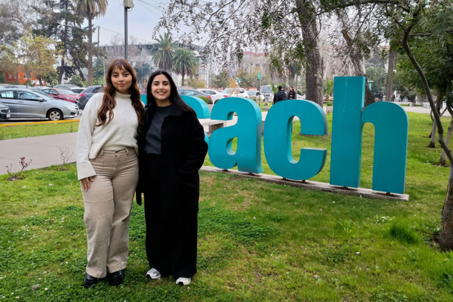 En la imagen aparecen las dos arquitectas  en los pastos de periodismo. Detrás, las letras monumentales USACH en verde corporativo.