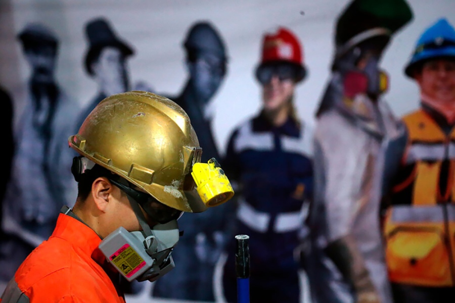 En primer plano aparece un minero con casco dorado y mascarilla de protección respiratoria exhibiendo el perfil derecho de su cuerpo. DE fondo, una gigantografía de trabajadores de la minería
