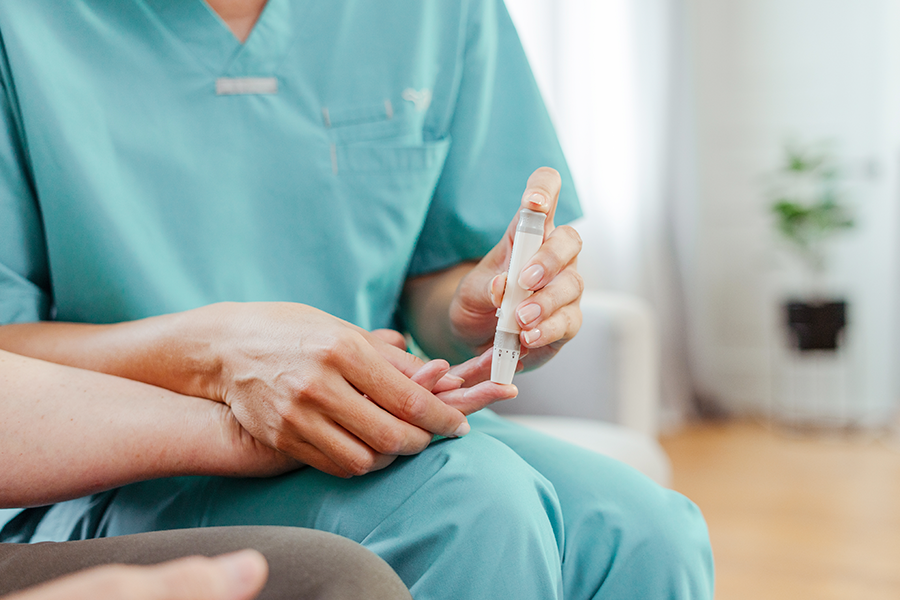 Imagen borrosa de un profesional sanitario con bata turquesa sosteniendo un dedo y un dispositivo médico, posiblemente una lanceta, para medir el nivel de azúcar en sangre.