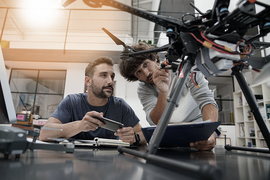 Imagen de dos personas en un taller, trabajando en un dron de gran tamaño. Uno sostiene un teléfono inteligente, mientras que el otro realiza ajustes en el dron.