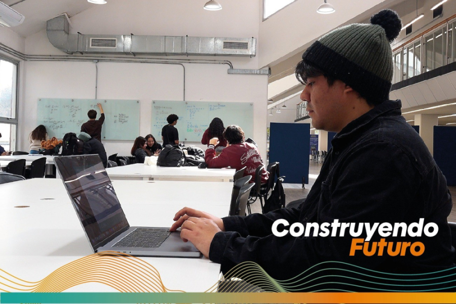 En la imagen aparece un estudiante de la universidad trabajando en su computador. Lleva un gorro de lana con un ponpon en su cabeza y está sentando a una mesa de la Biblioteca Central Usach