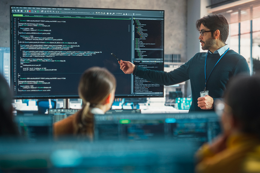 Um professor assinala uma grande tela, na qual são exibidas linhas de código de computador, durante uma aula em um ambiente educacional tecnológico.