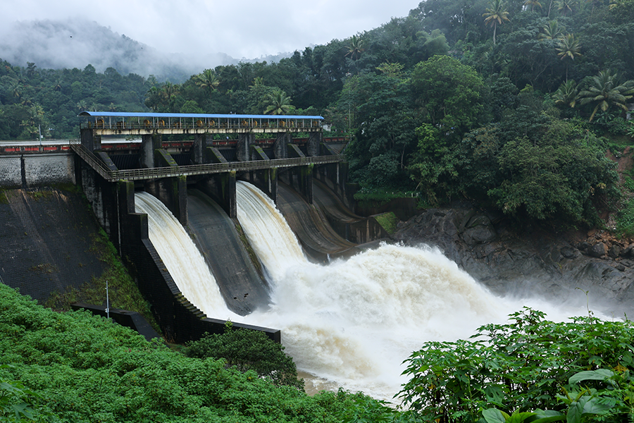 Uma barragem de concreto em uma floresta tropical tem três comportas abertas que liberam potentes cachoeiras de água branca em direção a um rio.