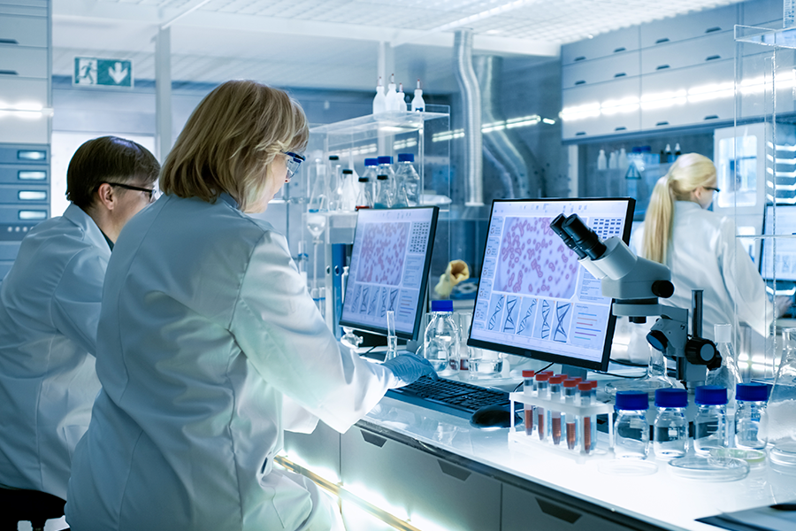 Cientistas com jalecos brancos trabalham na visualização de dados de células e DNA em um laboratório luminoso e moderno, rodeados por microscópios e tubos de ensaio.
